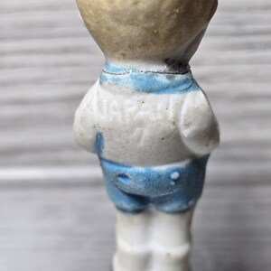 Puede incluir: Figura vintage de un ni&ntilde;o peque&ntilde;o, vista desde atr&aacute;s. La figura viste una camisa blanca con cuello azul y pantalones cortos azules. La figura est&aacute; hecha de un material de color claro, posiblemente porcelana o cer&aacute;mica.