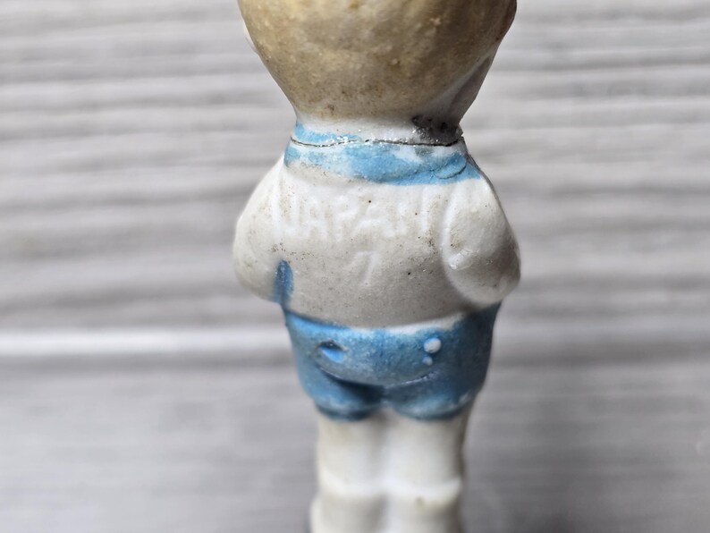 Puede incluir: Figura vintage de un ni&ntilde;o peque&ntilde;o, vista desde atr&aacute;s. La figura viste una camisa blanca con cuello azul y pantalones cortos azules. La figura est&aacute; hecha de un material de color claro, posiblemente porcelana o cer&aacute;mica.