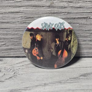 May include: A round button featuring the band Stray Cats. The button has a white border with a red zig-zag design. The image on the button shows the band members with their instruments. The band's name is printed in white letters.