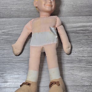 May include: A vintage doll with a plastic head and soft body. The doll has blonde hair, blue eyes, and is wearing a pink shirt and pants with a white diaper-like section. It has tan shoes with brown bows.