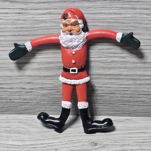 May include: A red Santa Claus figurine with a white beard, hat trim, and cuffs. The figure has black boots and gloves, a black belt, and a white fur trim on the coat. The arms and legs are poseable.