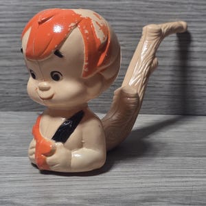 May include: A vintage plastic toy figure of a cartoon character with orange hair and a tan body. The figure is holding a black object and has a tan, curved tail. The toy is set against a gray wooden background.