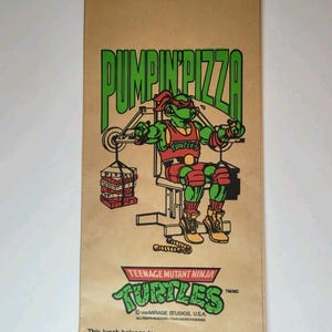 May include: Brown paper lunch bag featuring a Teenage Mutant Ninja Turtle character lifting weights, with the text "PUMPIN' PIZZA" in green. The bag also includes the text "TEENAGE MUTANT NINJA TURTLES".