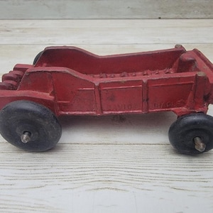 May include: A vintage red toy truck with black wheels. The truck has a rectangular bed and a cab with a detailed front end. The wheels are black and appear to be made of rubber. The toy is likely made of cast iron.