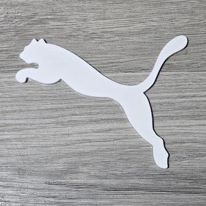 Op de afbeelding: Een wit Puma-logo in de vorm van een springende poema. Het logo is een strak, schoon ontwerp, tegen een grijze houtnerf achtergrond. De poema wordt in volle sprong afgebeeld, met uitgestrekte staart.