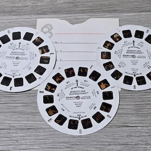 Könnte beinhalten: Drei weiße, kreisförmige View-Master-Scheiben mit Wrestling Superstars-Bildern. Jede Scheibe enthält sieben Paare kleiner fotografischer Dias mit aufgedruckten Textbeschriftungen wie Wrestling Superstars, 004-858 und 4067, zusammen mit einer weißen Papierhülle.