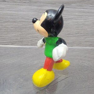 Pode incluir: Uma pequena figura de pl&aacute;stico do Mickey Mouse vestindo uma camisa verde, cal&ccedil;as vermelhas e sapatos amarelos. A figura est&aacute; de p&eacute; com a perna direita para a frente e o bra&ccedil;o esquerdo levantado.
