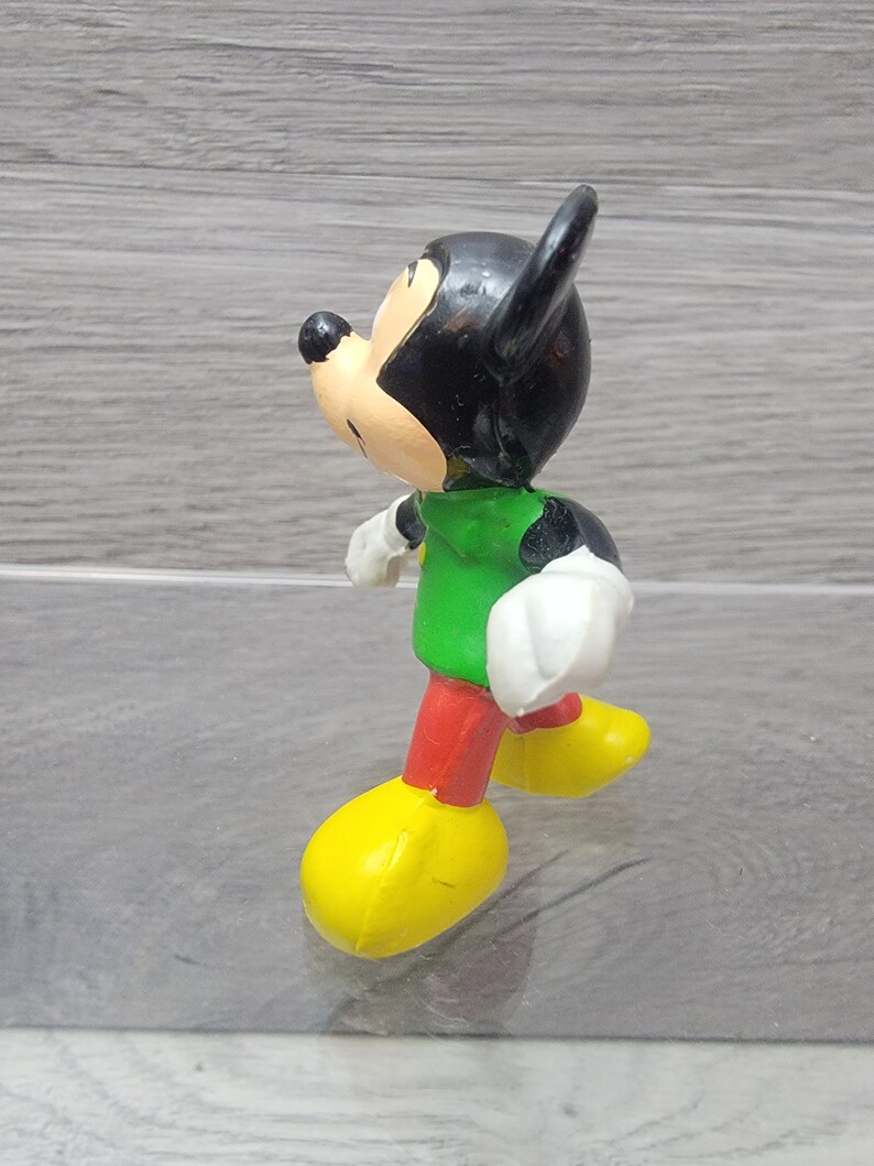 Pode incluir: Uma pequena figura de pl&aacute;stico do Mickey Mouse vestindo uma camisa verde, cal&ccedil;as vermelhas e sapatos amarelos. A figura est&aacute; de p&eacute; com a perna direita para a frente e o bra&ccedil;o esquerdo levantado.