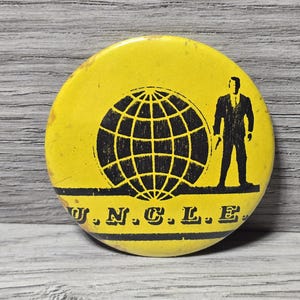 Könnte beinhalten: Gelber Knopf mit schwarzem Globus-Grafik und einer Silhouette eines Mannes im Anzug. Der Text "U.N.C.L.E." ist unten in Schwarz gedruckt. Der Knopf ist rund und weist leichte Gebrauchsspuren auf.