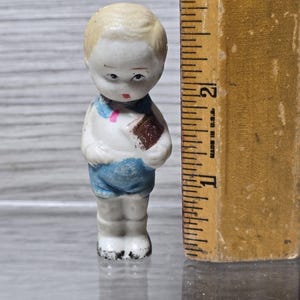 Puede incluir: Peque&ntilde;a figura de porcelana vintage de un ni&ntilde;o, sosteniendo un libro. La figura est&aacute; pintada con una camisa blanca, pantalones cortos azules y cabello rubio. La figura mide aproximadamente 5 cm de alto.