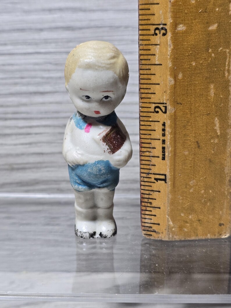 Puede incluir: Peque&ntilde;a figura de porcelana vintage de un ni&ntilde;o, sosteniendo un libro. La figura est&aacute; pintada con una camisa blanca, pantalones cortos azules y cabello rubio. La figura mide aproximadamente 5 cm de alto.