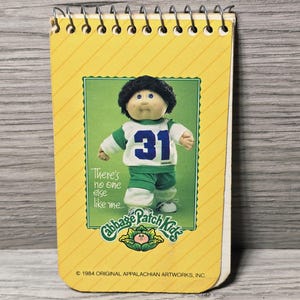 May include: A spiral-bound notepad with a yellow cover featuring a Cabbage Patch Kids graphic. The graphic shows a doll wearing a white and green jersey with the number 31. The notepad has the text "There's no one else like me..." and the Cabbage Patch Kids logo.