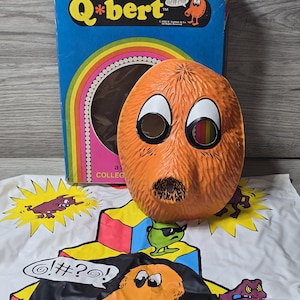 Può includere: Una maschera arancione di Q*bert con grandi occhi bianchi e baffi neri è posta davanti a una scatola blu con il logo Q*bert. Un tappetino da gioco bianco con personaggi di Q*bert è in primo piano. La maschera e la scatola sono oggetti da collezione vintage.