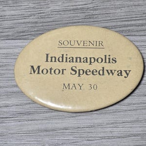 May include: An oval-shaped beige pinback button featuring black text that reads SOUVENIR, Indianapolis Motor Speedway, and MAY 30. The button has a smooth, glossy finish and is designed to be worn as a collectible accessory or memorabilia item.