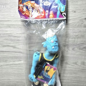 May include: A blue plush toy of a Space Jam character, in a clear plastic bag. The figure wears a black and yellow basketball uniform and trainers. The packaging includes a "Tune Squad" header and warning text.
