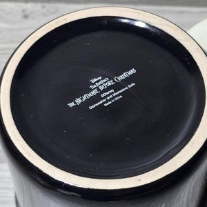 Puede incluir: Taza de cer&aacute;mica negra con asa color crema. La base de la taza presenta el texto "Tim Burton's The Nightmare Before Christmas" y "&copy;Disney". La taza es apta para lavavajillas y microondas.