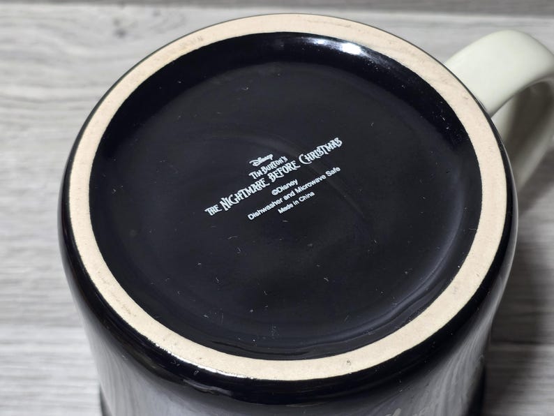 Puede incluir: Taza de cer&aacute;mica negra con asa color crema. La base de la taza presenta el texto "Tim Burton's The Nightmare Before Christmas" y "&copy;Disney". La taza es apta para lavavajillas y microondas.