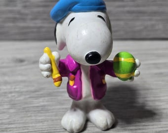 May include: A Snoopy figurine wearing a blue beret and a purple jacket, holding a yellow paintbrush and a green and yellow Easter egg. The figurine is white with black ears and a black nose.