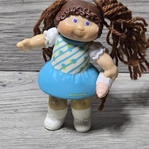 May include: A vintage Cabbage Patch Kids doll with brown yarn pigtails, blue eyes, and a rosy complexion. The doll wears a blue dress with a white and blue striped pattern and white socks. The doll is holding a pink object.