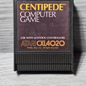 Puede incluir: Un cartucho de videojuego Atari Centipede vintage. La etiqueta presenta el título del juego en amarillo y blanco, con el logotipo de Atari y la información de copyright. El cartucho es negro.