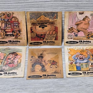 Op de afbeelding: Een set van tien vintage verzamelkaarten met cartoonillustraties. De kaarten zijn bruin met witte randen en bevatten de tekst "CB Jeebics" en "Sunbeam". De illustraties tonen verschillende scènes, waaronder een postkoets, een beer, een rolschaats, een auto, een cowboy en een man met een fles lotion.
