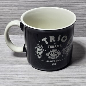 Puede incluir: Taza de cer&aacute;mica negra con interior y asa blancos. La taza presenta el texto "TRIO OF TERROR" e ilustraciones de personajes de una pel&iacute;cula. Las palabras "OOGIE'S CREW" tambi&eacute;n est&aacute;n impresas en la taza.