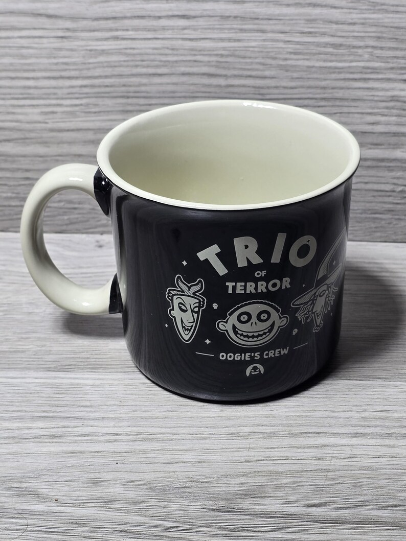 Puede incluir: Taza de cer&aacute;mica negra con interior y asa blancos. La taza presenta el texto "TRIO OF TERROR" e ilustraciones de personajes de una pel&iacute;cula. Las palabras "OOGIE'S CREW" tambi&eacute;n est&aacute;n impresas en la taza.