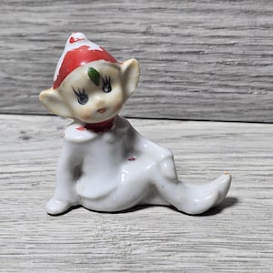 Puede incluir: Pequeña figura de elfo de cerámica. El elfo es blanco con un gorro a rayas rojas y blancas y un cuello rojo. El elfo está sentado con las piernas extendidas. La figura tiene orejas grandes y una expresión dulce.