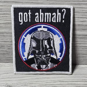 以下が含まれることがあります： 黒い正方形のパッチ。白い縁取りがあり、青と赤の円の中にダース・ベイダーのヘルメットが描かれています。「got ahmah?」の文字が画像の上にあります。パッチはアイロン接着または縫い付け用です。