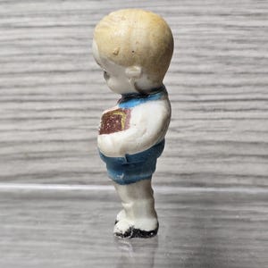 Puede incluir: Peque&ntilde;a figura vintage de un ni&ntilde;o, vista de perfil. La figura tiene una cabeza rubia, una camisa blanca, pantalones cortos azules y zapatos negros. El ni&ntilde;o sostiene un libro con una cubierta marr&oacute;n.