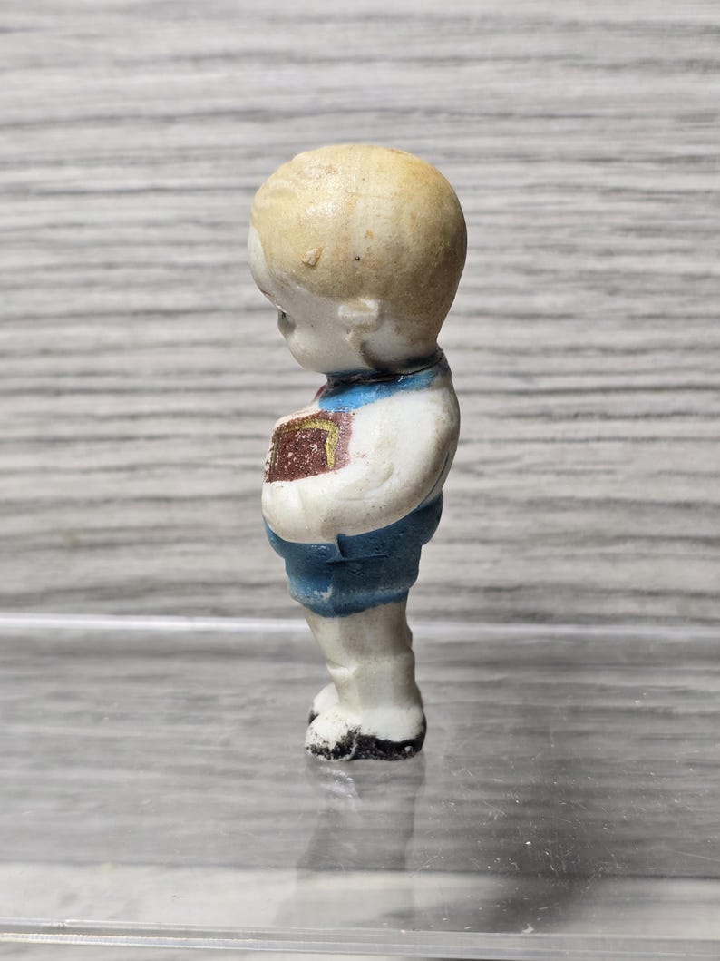 Puede incluir: Peque&ntilde;a figura vintage de un ni&ntilde;o, vista de perfil. La figura tiene una cabeza rubia, una camisa blanca, pantalones cortos azules y zapatos negros. El ni&ntilde;o sostiene un libro con una cubierta marr&oacute;n.