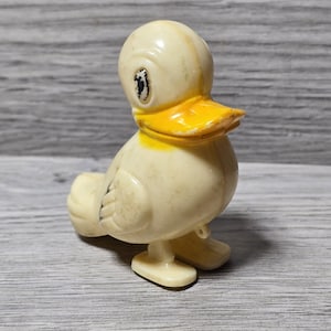 May include: A vintage, off-white plastic duck toy with a bright yellow beak. The duck has black eyes and is standing on two small feet. The toy has a classic, cartoonish design.