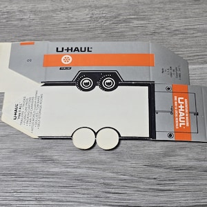 May include: A folded U-Haul cardboard box featuring a trailer design. The box is grey and white with orange accents and the U-Haul logo. The text "U-HAUL" and "Mas it ALL" are visible.