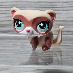 May include: A tan and brown ferret figurine with large, teal eyes and black eyelashes. The toy ferret has a white face and pink inner ears. Its pose is crouching, with a curved tail.