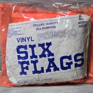 Könnte beinhalten: Ein verpackter Vinyl-Poncho in einer orangefarbenen Plastiktüte. Das weiße Etikett trägt den blauen Schriftzug "SIX FLAGS" und "VINYL PONCHO". Das Etikett weist auch "100% WATERPROOF" aus. Der Poncho misst ca. 0 cm x 203 cm.