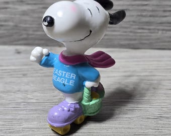 May include: A Snoopy figurine in a blue shirt with the text "Easter Beagle", a purple scarf, and purple roller skates. Snoopy is holding a green basket. The figurine is on a wooden surface.