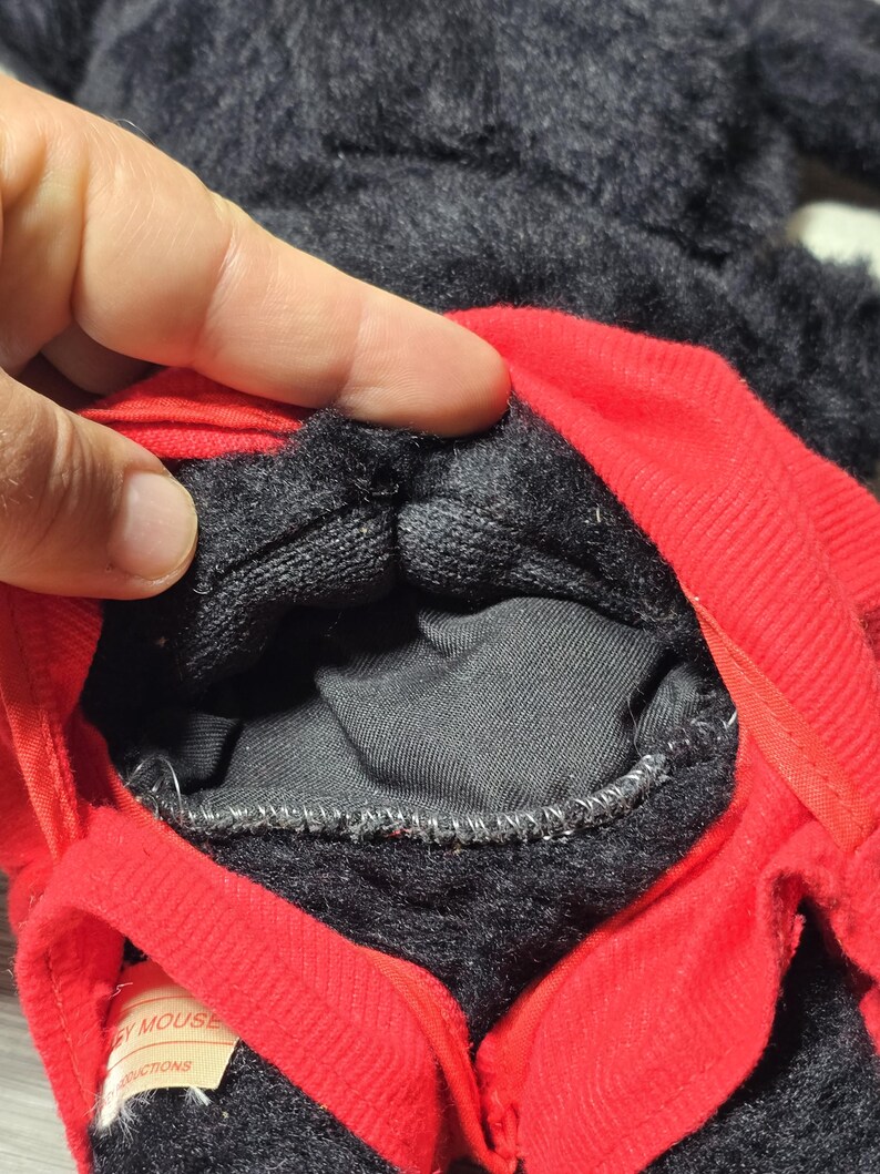 May include: Close-up of the inside of a black and red Mickey Mouse plush toy. The toy has a zipper and a label that reads "Mickey Mouse" and "Instructions".