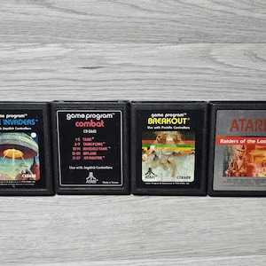 Puede incluir: Cuatro cartuchos de juego Atari 2600 negros alineados horizontalmente. Las etiquetas muestran los títulos Space Invaders, Combat, Breakout y Raiders of the Lost Ark, cada uno con arte de portada único y texto que detalla el juego y los mandos.