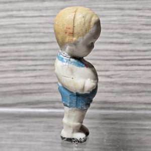 Puede incluir: Peque&ntilde;a figura vintage de un ni&ntilde;o. La figura tiene una cabeza rubia, pantalones cortos azules y una camisa blanca con rayas azules. La figura est&aacute; de pie y parece estar hecha de cer&aacute;mica o porcelana.