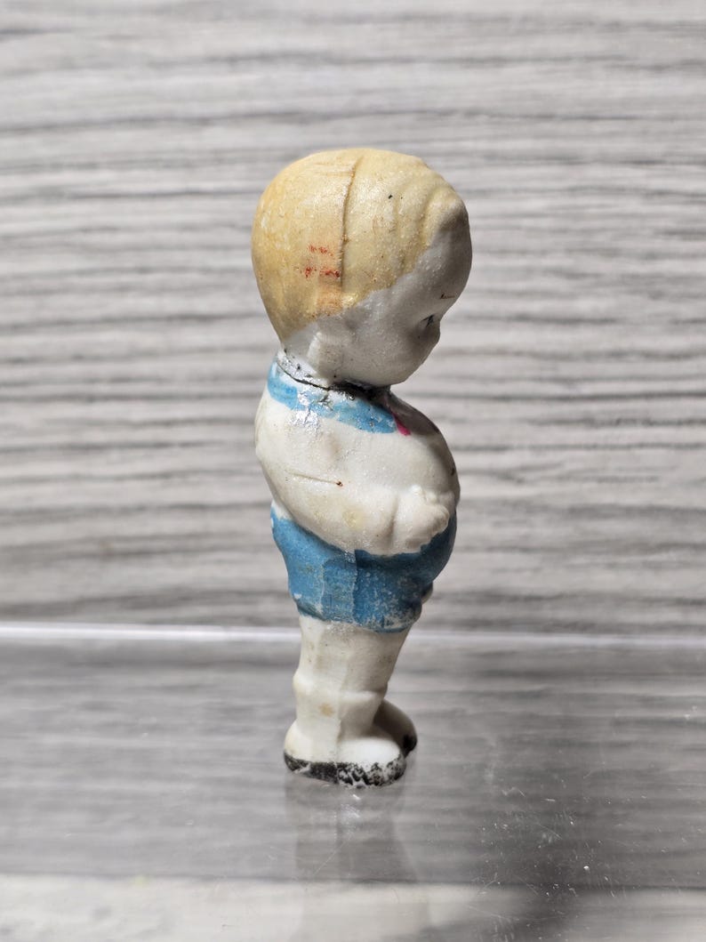 Puede incluir: Peque&ntilde;a figura vintage de un ni&ntilde;o. La figura tiene una cabeza rubia, pantalones cortos azules y una camisa blanca con rayas azules. La figura est&aacute; de pie y parece estar hecha de cer&aacute;mica o porcelana.