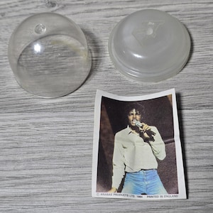 May include: Clear plastic dome and lid with a sticker of a singer holding a microphone. The sticker has the text "ANABAS PRODUCTS LTD. 1984 PRINTED IN ENGLAND". The singer is wearing a light-colored shirt and blue jeans.