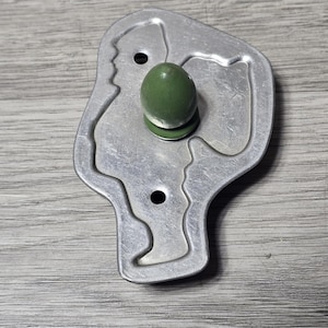 May include: A silver-colored metal cookie cutter shaped like a profile of a person wearing a hat. The cutter features a smooth, rounded green handle attached to the center, and two small circular holes are visible within the metal frame.