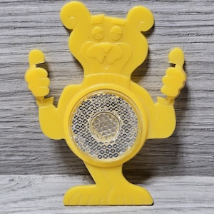 May include: Yellow plastic bear-shaped bicycle reflector with a reflective silver circle in the centre. The bear is smiling and has its arms raised in the air.