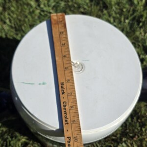 May include: White plastic lid with a metal ring handle. A wooden ruler with the text "Buick - Chevrolet - Phone 21" is next to the lid.