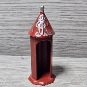 May include: A small, cylindrical red metal structure with an open front and a conical roof. The roof is decorated with silver-colored painted scrollwork and topped with a small silver sphere. The base is hexagonal, and the overall design resembles a miniature tower or a decorative architectural element.