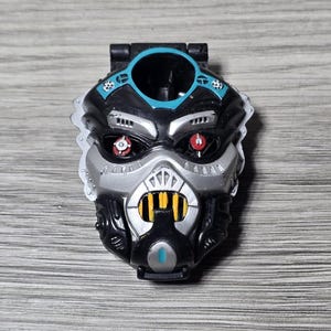 Può includere: Un volto di robot giocattolo nero, argento e verde acqua con occhi rossi e dettagli gialli. Il giocattolo ha una parte superiore incernierata e un'apertura circolare. Il design è dettagliato con elementi meccanici e futuristici.