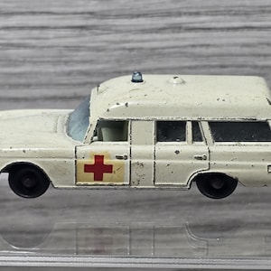 May include: A vintage off-white die-cast toy ambulance with black wheels, a small blue light on the roof, and a red cross symbol on the front door. The paint shows signs of wear and chipping, with a clear windshield and side windows.
