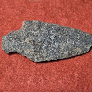Puede incluir: Una piedra gris de forma irregular con una textura rugosa. La piedra está sobre un fondo rojo. La piedra parece ser un artefacto natural, posiblemente una herramienta o un trozo de materia prima.