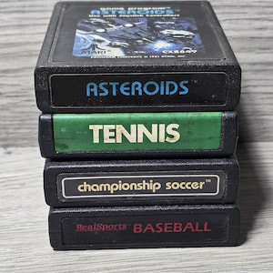 May include: A stack of four black plastic Atari video game cartridges. The top cartridge features an Asteroids label with blue text. Below are cartridges labeled Tennis in white on green, Championship Soccer in white, and RealSports Baseball in red.