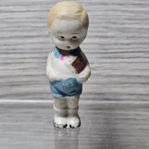 Puede incluir: Peque&ntilde;a figura de porcelana vintage de un ni&ntilde;o. La figura est&aacute; pintada con una camisa blanca, pantalones cortos azules y un libro marr&oacute;n. El ni&ntilde;o tiene el pelo rubio y una expresi&oacute;n ligeramente triste. La figura est&aacute; de pie.